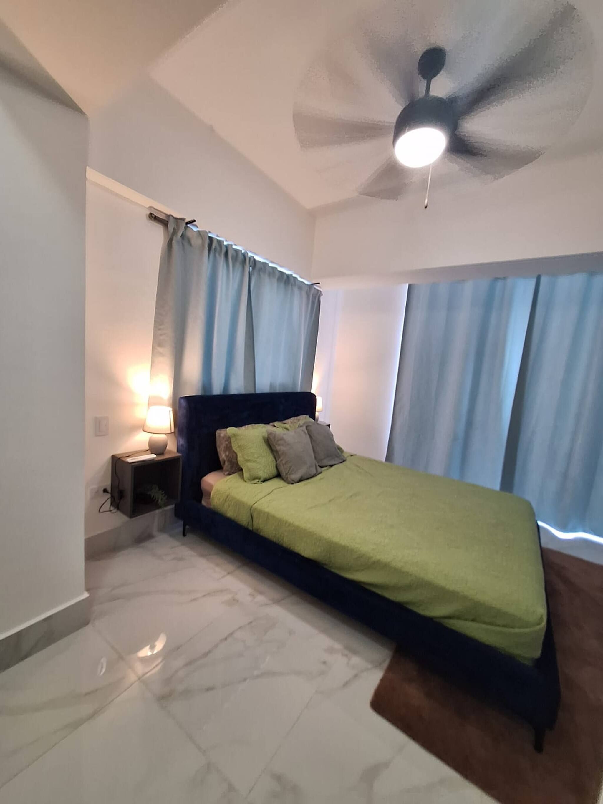 Une chambre à coucher avec un lit, une table de chevet, une lampe, un ventilateur de plafond et des rideaux.