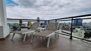 En balkon med to liggestole og et bord, der udover en byudsigt med bygninger og en overskyet himmel.