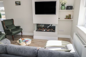 A living room with a fireplace, a television mounted on the wall, a green armchair, and a grey sofa. - RED HOUSE, pet friendly, with open fire in Carraroe, County Galway (Galway)