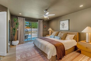 A bedroom with a bed, ceiling fan, bedside lamps, and a view of the outdoors.