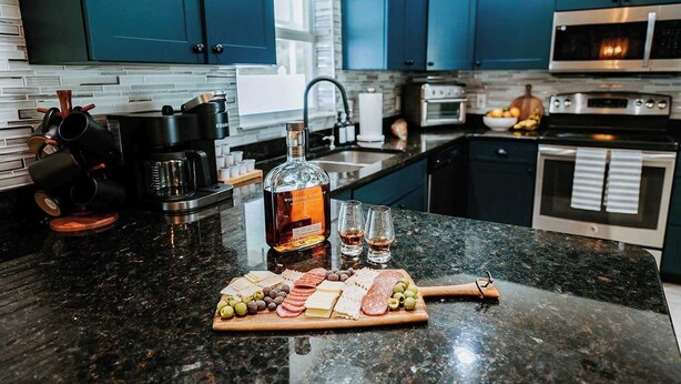 A kitchen with dark blue cabinets, a granite countertop, a coffee machine, a bottle of liquor, glasses, and a charcuterie board. - Sleeps 10 • Outdoor Oasis • Heated Pool • Grill (Bradenton)