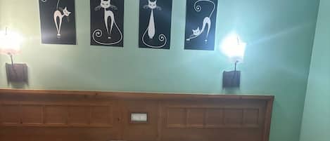 Una cama bien hecha con una colcha estampada, dos mesitas de noche con lámparas, y cuatro cuadros con motivos de gatos colgados en la pared.