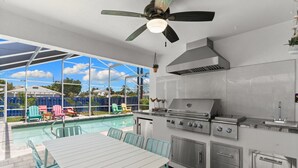 Une cuisine extérieure équipée d’un barbecue, d’une table à manger avec des chaises, et un espace piscine visible à travers une paroi vitrée.