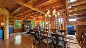 A wooden interior with a dining table set for four, a fireplace, and a large window overlooking greenery. - Little Bear: 3-bedroom cabin with Hot Tub, Putt Putt, Game Room & Fire Pit (Sevierville)