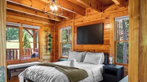 A wooden cabin bedroom with a bed, bedside tables, a bench, and large windows. - Little Bear: 3-bedroom cabin with Hot Tub, Putt Putt, Game Room & Fire Pit (Sevierville)