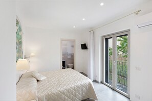 Ein Schlafzimmer mit einem Bett, einem Nachttisch, einer Wandlampe, einer Glastür zu einem Balkon und einem durch eine offene Tür einsehbaren Badezimmer.