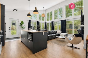 A modern kitchen with a central island, a dining area with a sofa and coffee table, and large windows with a view of greenery. - Private Sunken Spa & Sunroom - Luxury Chalet (Pont-Rouge)