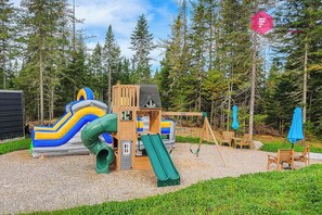 Un parc infantil amb tobogans i cadira d'oscalls envoltat d'arbres.