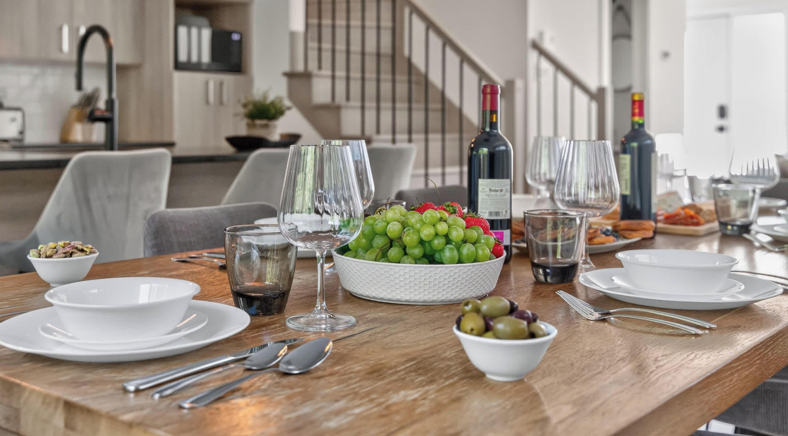 A dining table set with plates, cutlery, a bowl of grapes, a bowl of olives, wine glasses, and bottles.