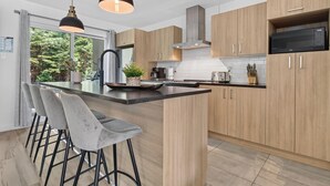 Moderni keittiö, jossa on puukalusteet, keskellä sijaitseva saareke baarituoleineen, sisäänrakennettu mikroaaltouuni ja suuri ikkuna, josta avautuu näkymä vehreään maisemaan.