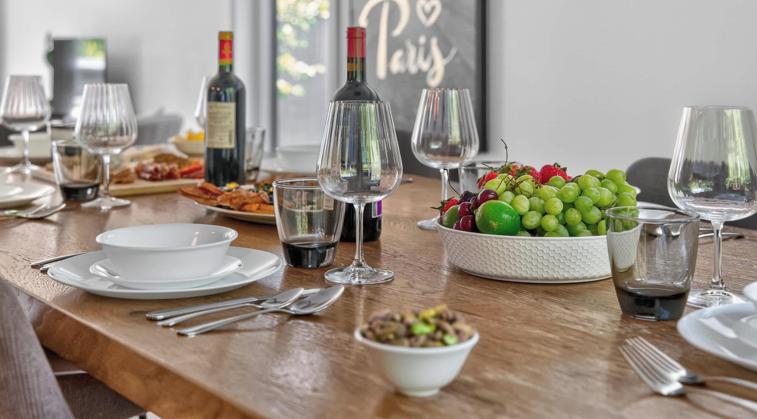Una mesa de comedor preparada con copas de vino, platos, cubiertos y un bol de fruta.