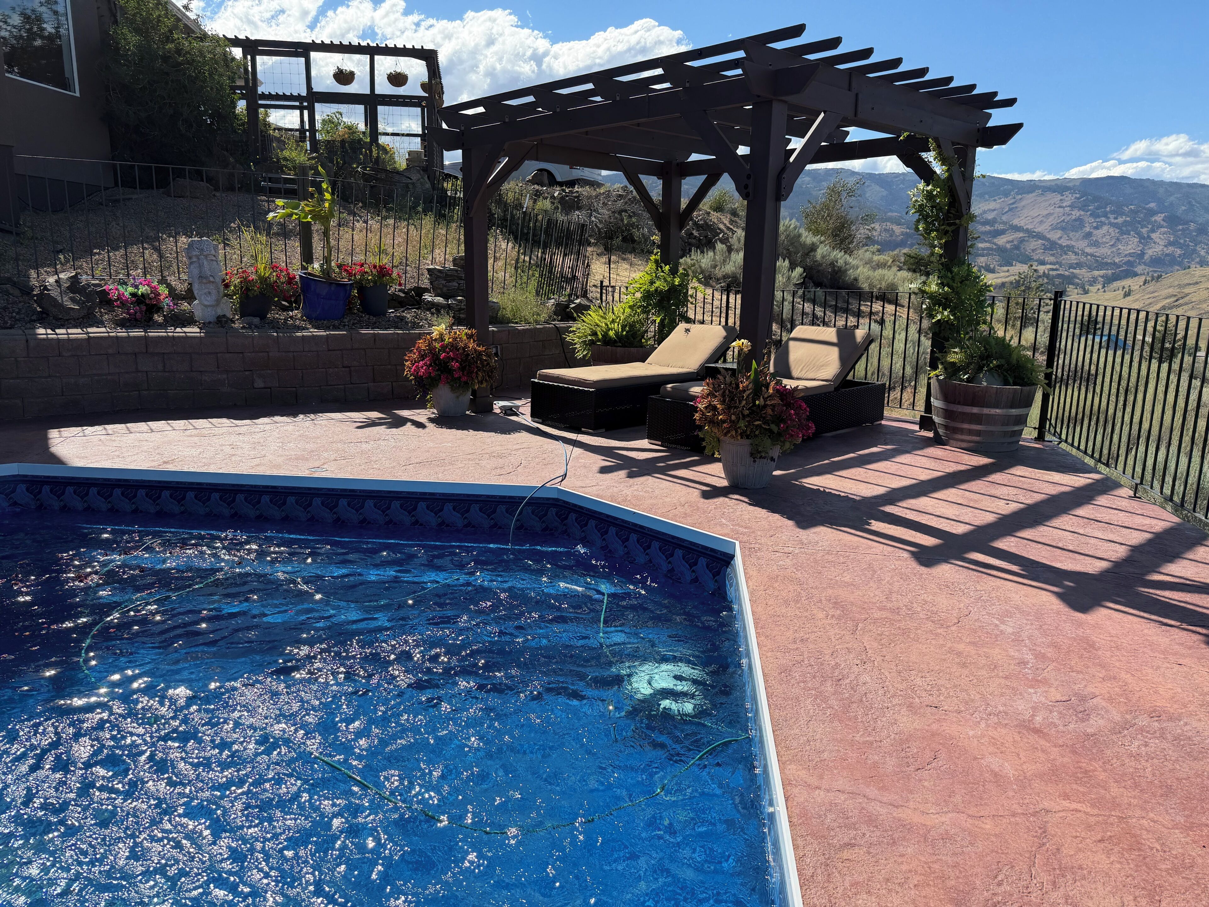 A swimming pool with a pergola and lounge chairs, surrounded by a fenced patio area with potted plants and a view of mountains.
