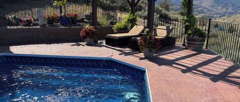 A swimming pool with a pergola and lounge chairs, surrounded by a fenced patio area with potted plants and a view of mountains.