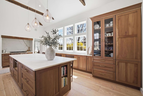 A spacious kitchen with wooden cabinets, a large island, and a window with a view of trees. - Historic Bones, Entirely New Soul | Designer Lux (Sandwich)
