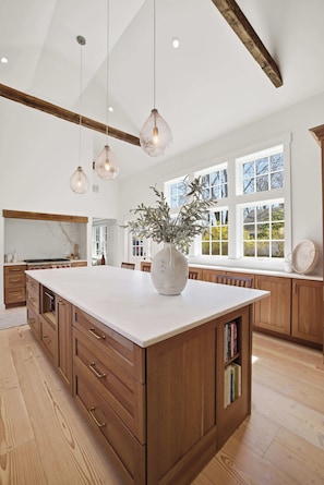 A spacious kitchen with a large island, wooden cabinetry, pendant lighting, and a view through large windows. - Historic Bones, Entirely New Soul | Designer Lux (Sandwich)