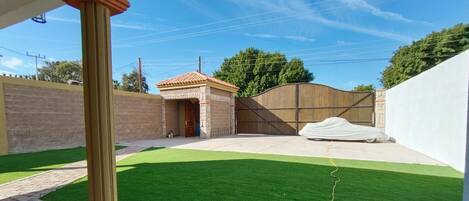 A fenced backyard with artificial grass, a covered car, and a building with a tiled roof.