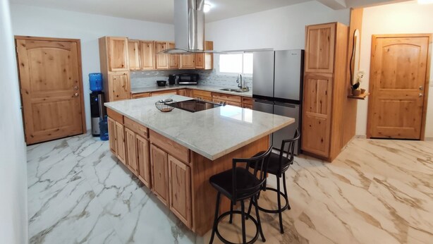 A modern kitchen with wooden cabinets, a stainless steel range hood, a double sink, and a large island with bar stools.