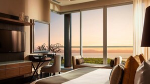 Ett hotellrum med en stor säng, ett skrivbord, en stol, en TV och utsikt över havet.