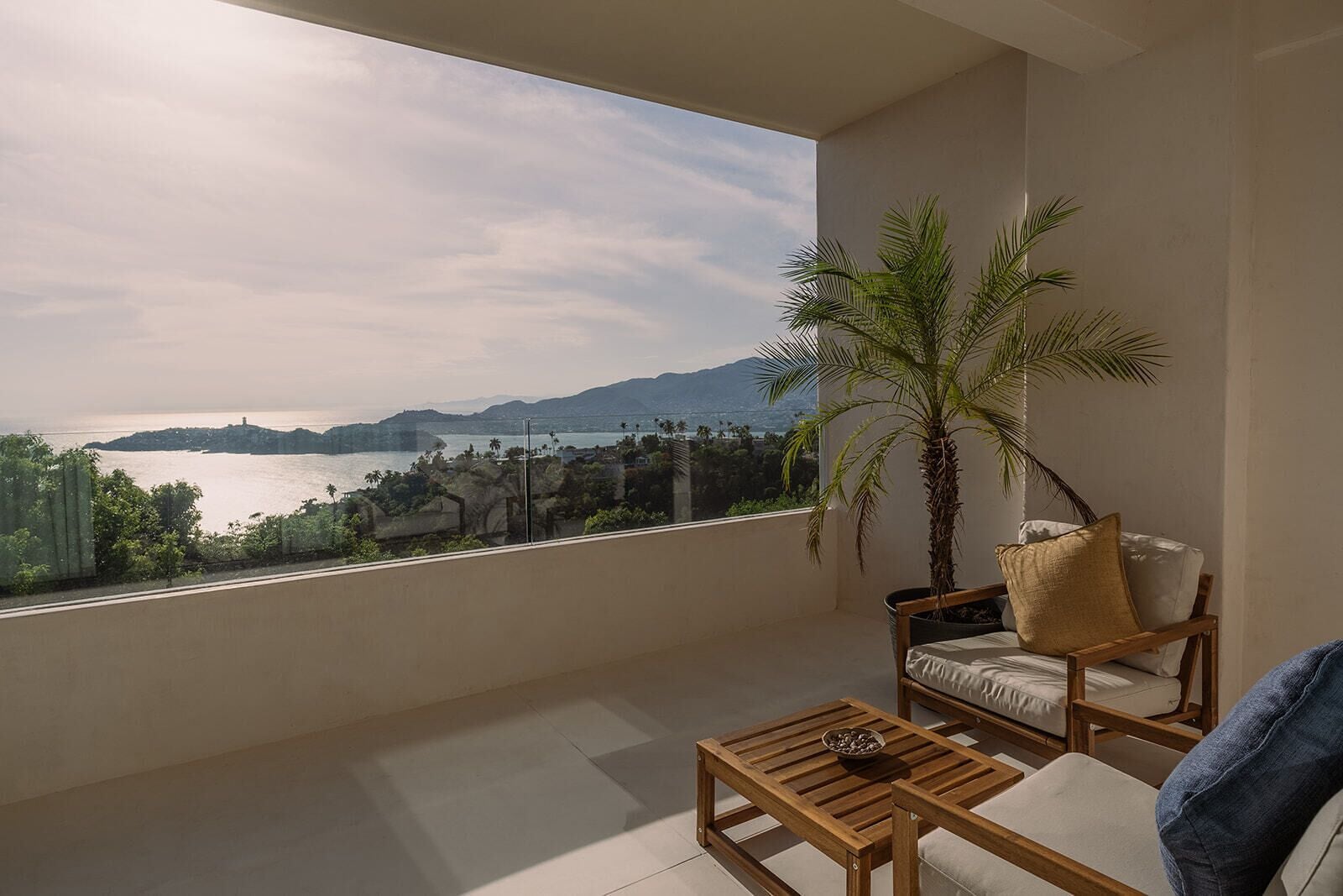 Un balcón con vista al agua y a las montañas, una mesa de madera, una silla y una palmera en maceta.