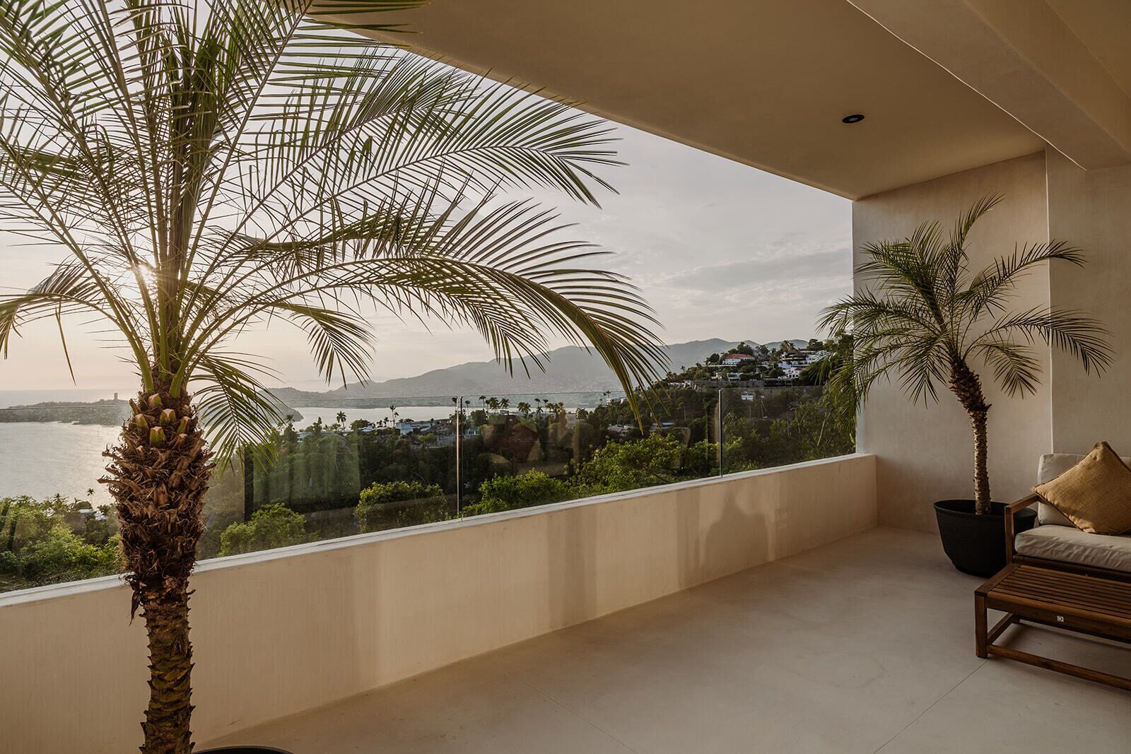 A balcony with a view of the sea and mountains, a palm tree, and a sofa with cushions.
