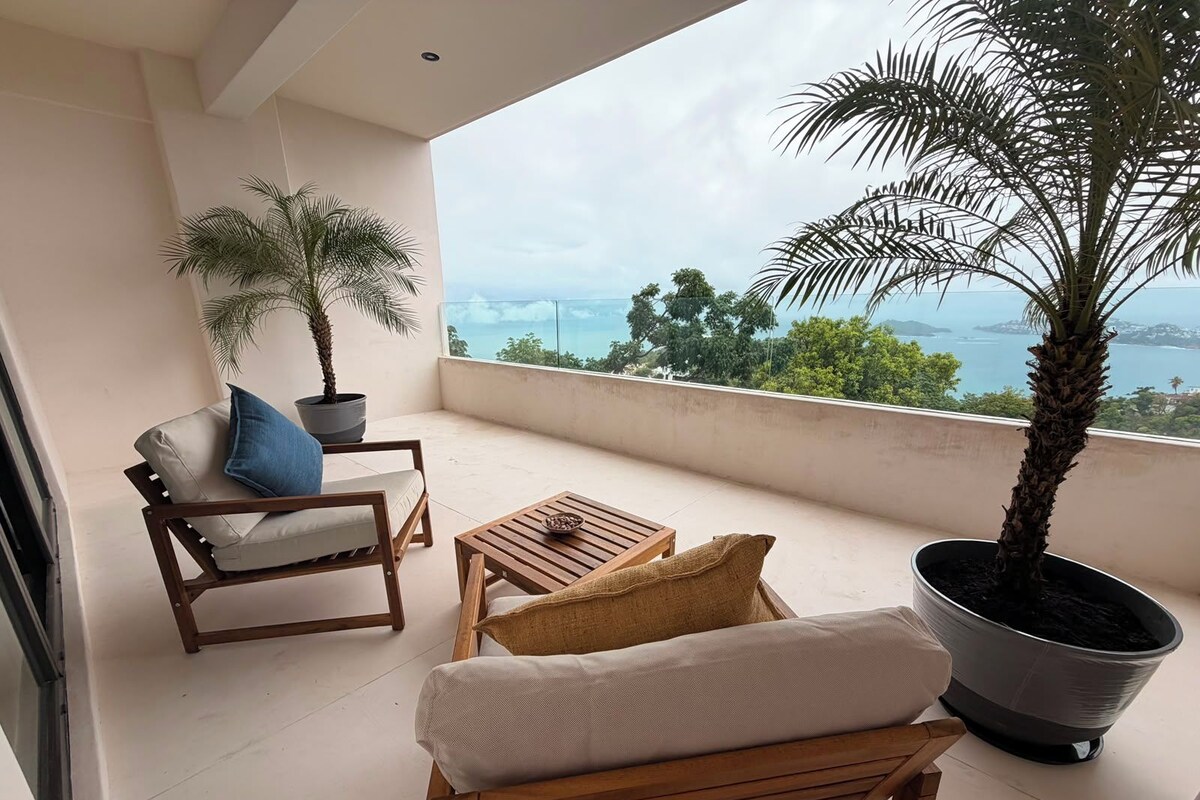 A balcony with a view of the sea, a wooden chair, a small table, and two potted plants.