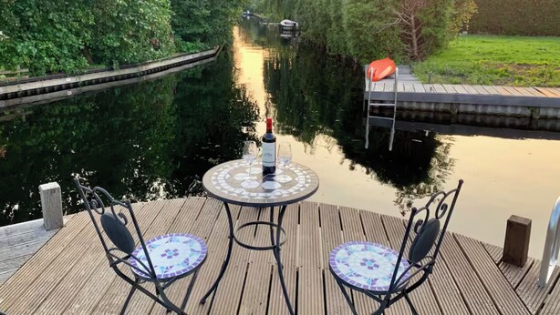 A wooden deck with two chairs and a table, a bottle of wine, and a view of a canal with a lock and a boat. - Sunny Pool Villa at Vinkeveense Plassen | 20 Min from Amsterdam (Vinkeveen)