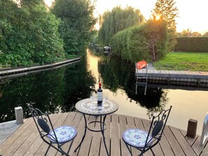 Een houten terras met twee stoelen, een tafel, een fles wijn en uitzicht op een gracht met een sluis en een boot.