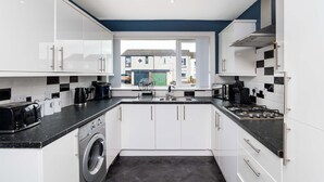 A modern kitchen with white cabinets, a built-in washing machine, and a view through the window. - Fife Coast Stay with Parking & Garden in Rosyth - WiFi (Rosyth)