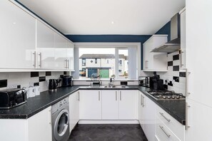 A modern kitchen with white cabinets, a built-in washing machine, and a view through the window. - Fife Coast Stay with Parking & Garden in Rosyth - WiFi (Rosyth)