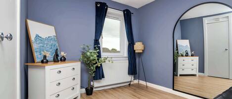 A bedroom with a white dresser, a potted plant, a window with curtains, a mirror, and a door.