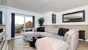 A modern living room with a sectional sofa, a coffee table, a large mirror, and a sliding glass door leading to a balcony. - Fife Coast Stay with Parking & Garden in Rosyth - WiFi (Rosyth)