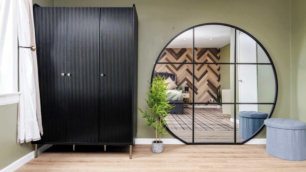 A room with a large black wardrobe, a round mirror, a potted plant, and a blue ottoman. - Fife Coast Stay with Parking & Garden in Rosyth - WiFi (Rosyth)