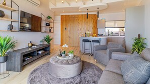 A modern living room with a grey sofa, a round ottoman, and a TV on a wall-mounted shelf. - Full spec apartment with stunning ocean views in the heart of Camps Bay (Cape Town)