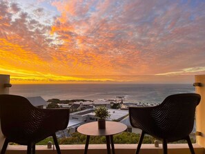 A balcony with two chairs and a small table, overlooking a coastal view with buildings and the sea. - Full spec apartment with stunning ocean views in the heart of Camps Bay (Cape Town)