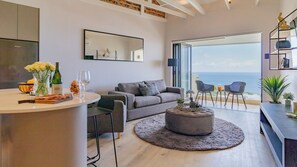 Un salon moderne avec un canapé gris, un pouf rond, une table à manger avec des chaises et une grande fenêtre donnant sur la mer.