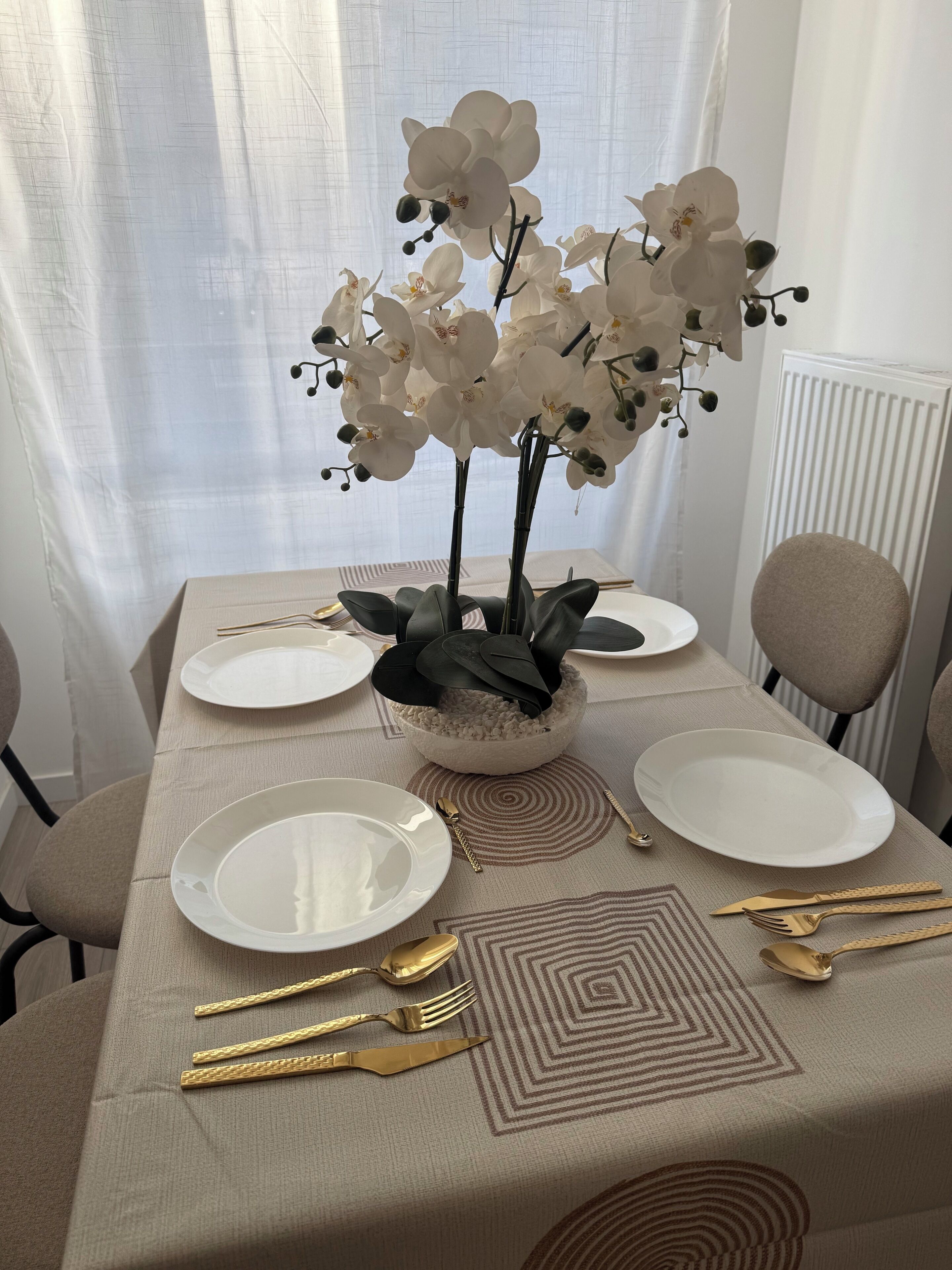 Een eettafel met witte borden, gouden bestek en een bloemstuk van witte orchideeën.