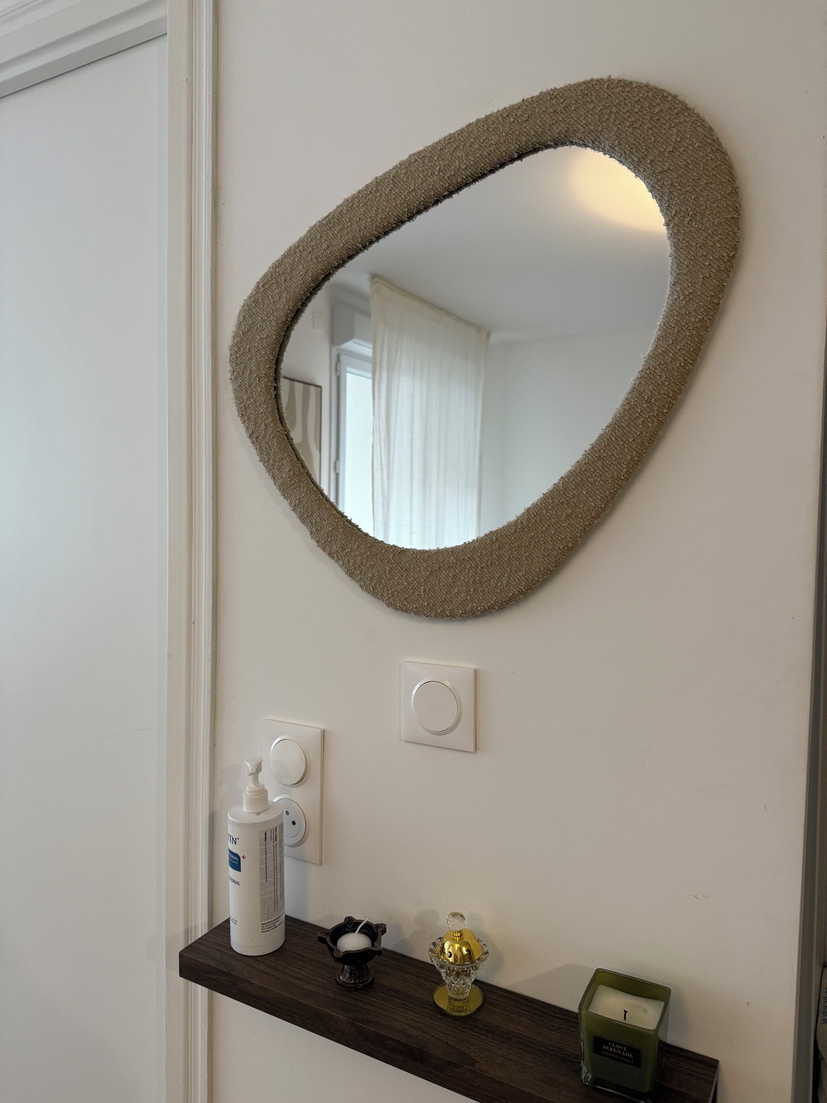 A mirror with a textured frame, a white door, a wall-mounted light switch, a shelf with a bottle, a candle, and a small decorative item.