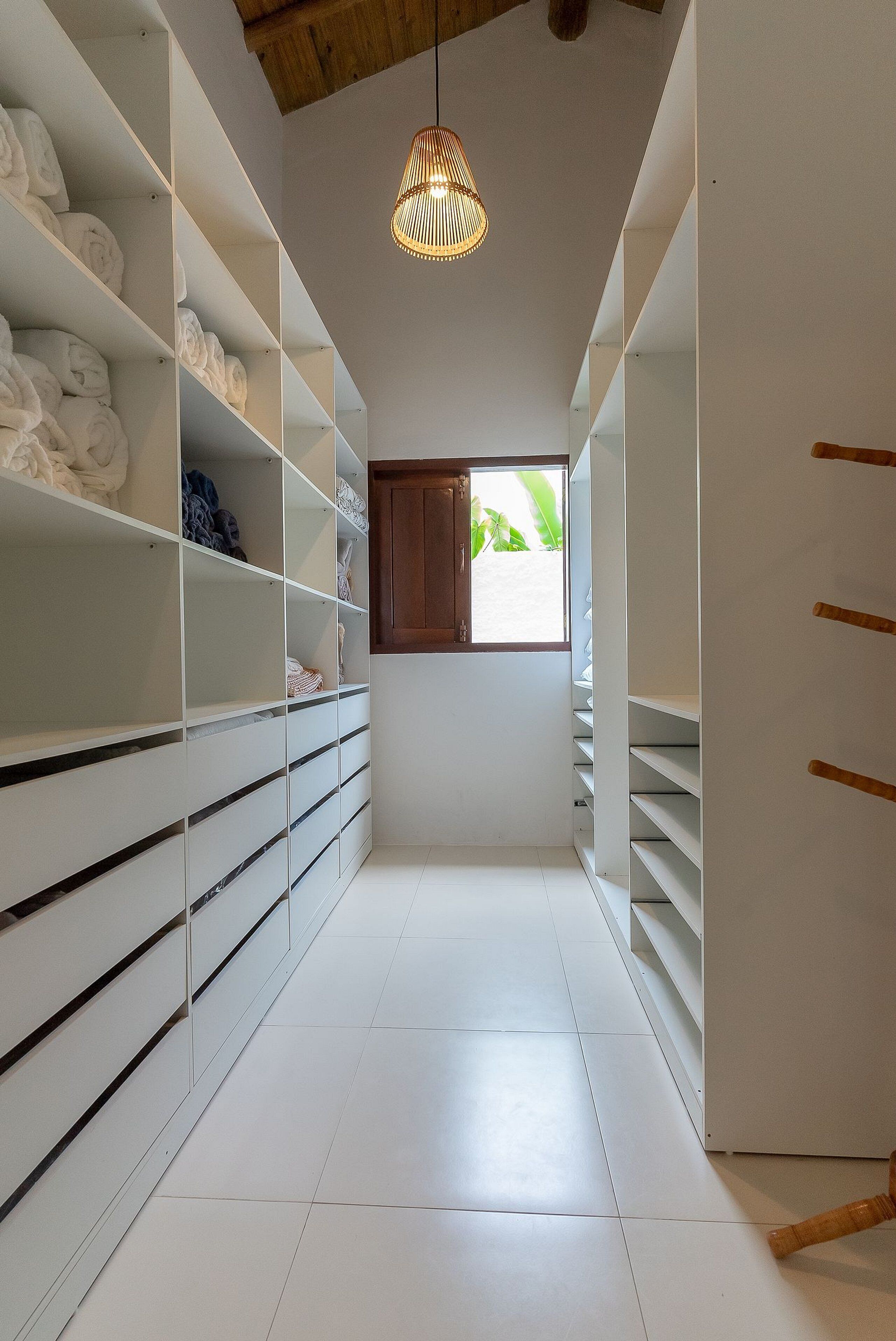 Um closet espaçoso com prateleiras e gavetas brancas, uma luminária pendente e uma janela com vista para o verde.