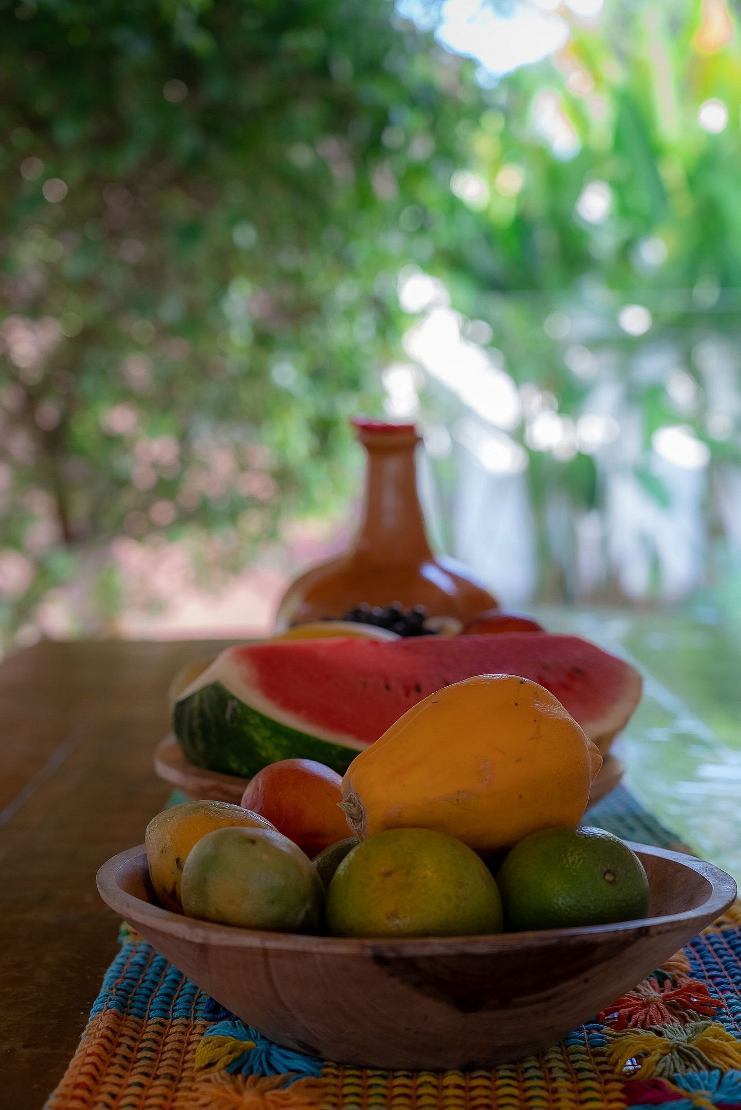 Un saladier en bois rempli de fruits et un pot en argile, le tout posé sur une nappe tissée.