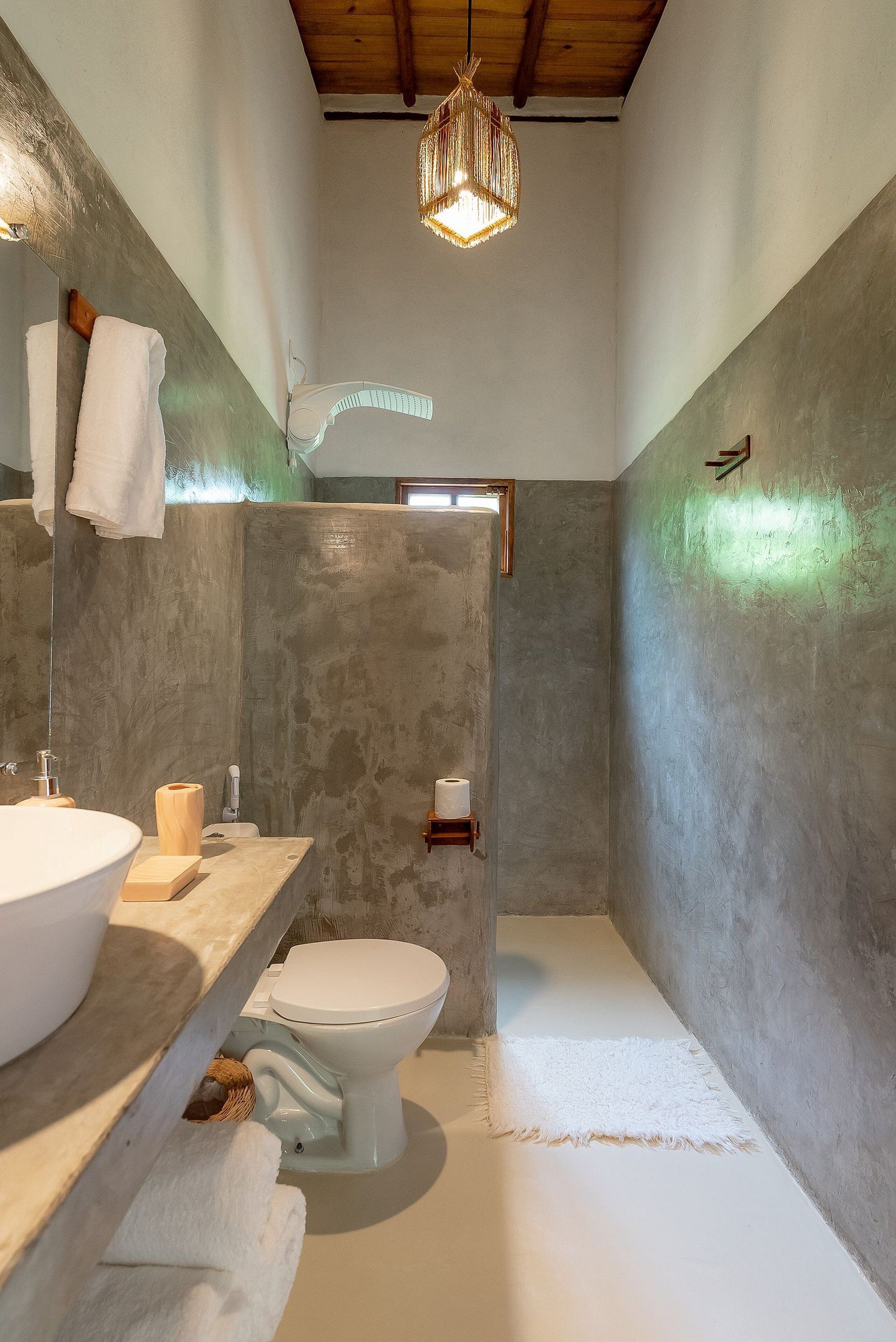 Une salle de bain dotée d’un grand lavabo, de toilettes et d’un espace douche, avec un mur en béton et un luminaire suspendu.
