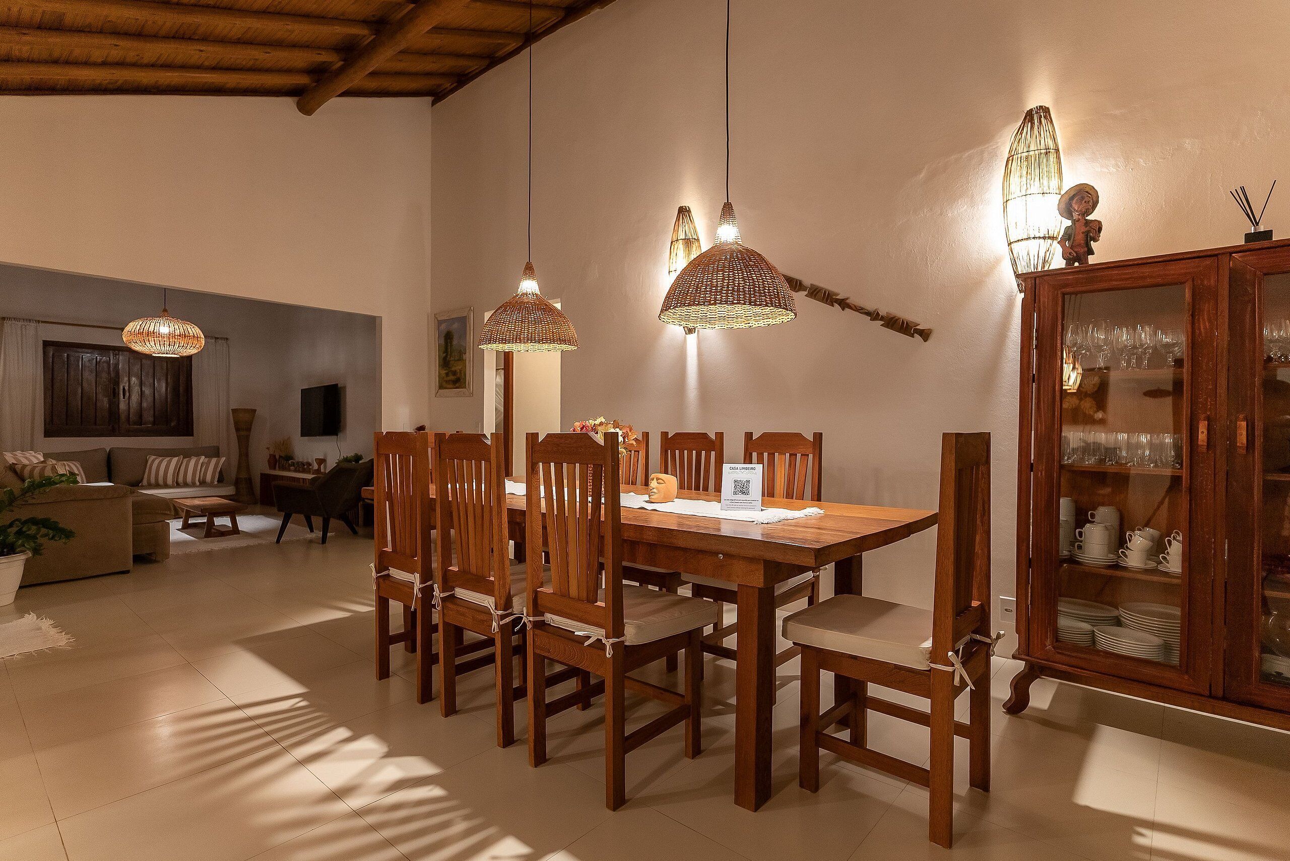 A dining area with a wooden table, chairs, pendant lights, and a cabinet with glassware.