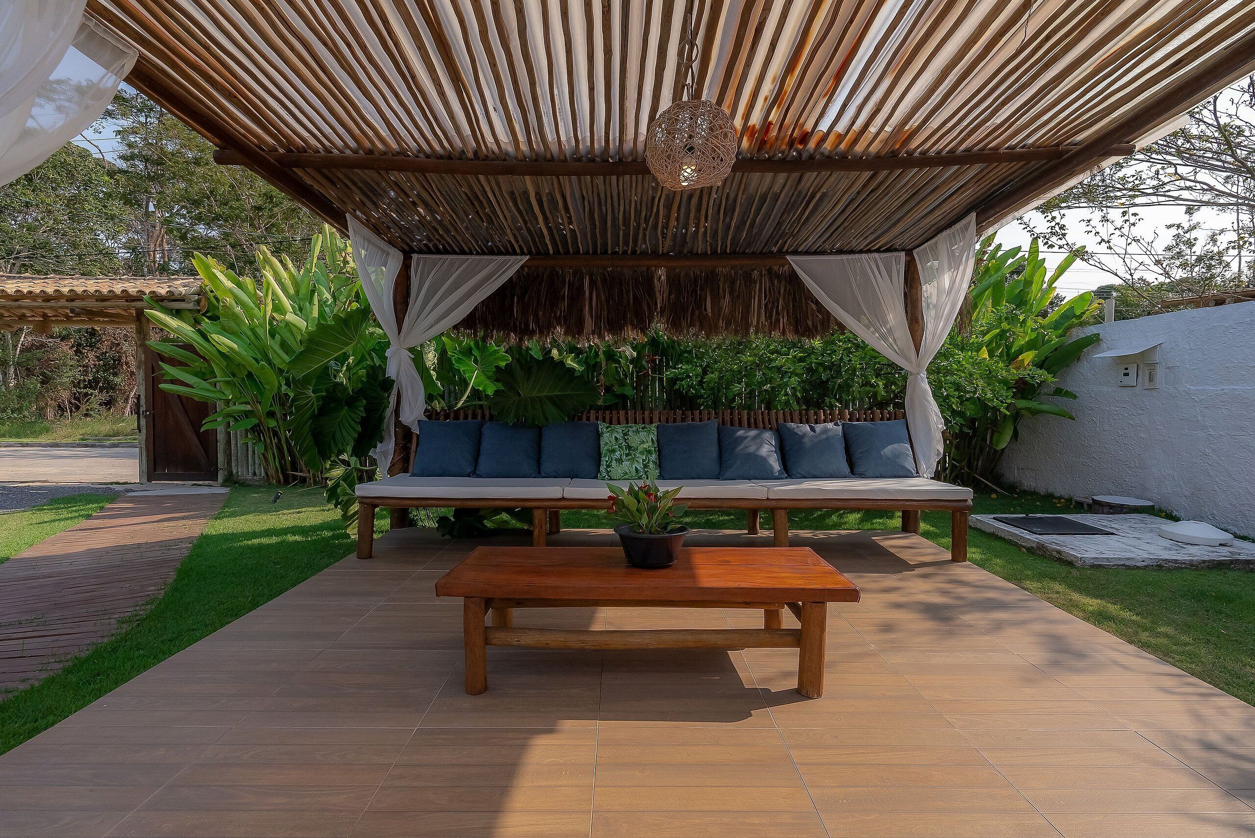 A covered outdoor seating area with a wooden table and a bench, surrounded by greenery.