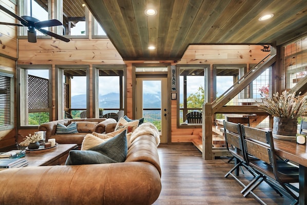Ein gemütliches Blockhaus-Interieur mit einem großen Ledersofa, einem Holzesstisch und einer Treppe, die auf einen Balkon mit Bergblick führt.