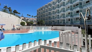 A swimming pool with a white fence, a multi-story building with balconies, and palm trees in the background. - Modern Premium Apartment Near Ccyumbo (San Bartolomé de Tirajana)