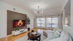 A modern living room with a flat-screen TV, a round coffee table, a sofa, and a large window with curtains. - 2 Homes • 8BR (sleeps 18) • Game Room & Backyards (Houston)