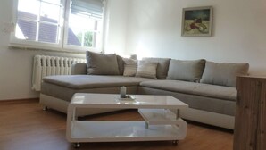 A living room with a sectional sofa, a glass coffee table, a radiator, and a window with blinds. - Ruhige Ferienwohnung Lachmöwe im Ostseebad Dranske auf Rügen (Dranske)