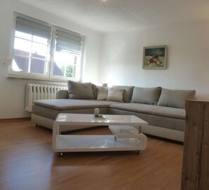 A living room with a sectional sofa, a glass coffee table, a radiator, and a window with blinds. - Ruhige Ferienwohnung Lachmöwe im Ostseebad Dranske auf Rügen (Dranske)