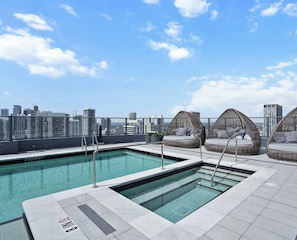 Ein Dachpool mit klarem, blauem Wasser, umgeben von weiß gefliestem Boden und einer Glasschutzwand. Drei Korbstühle mit Kissen sind vorhanden.