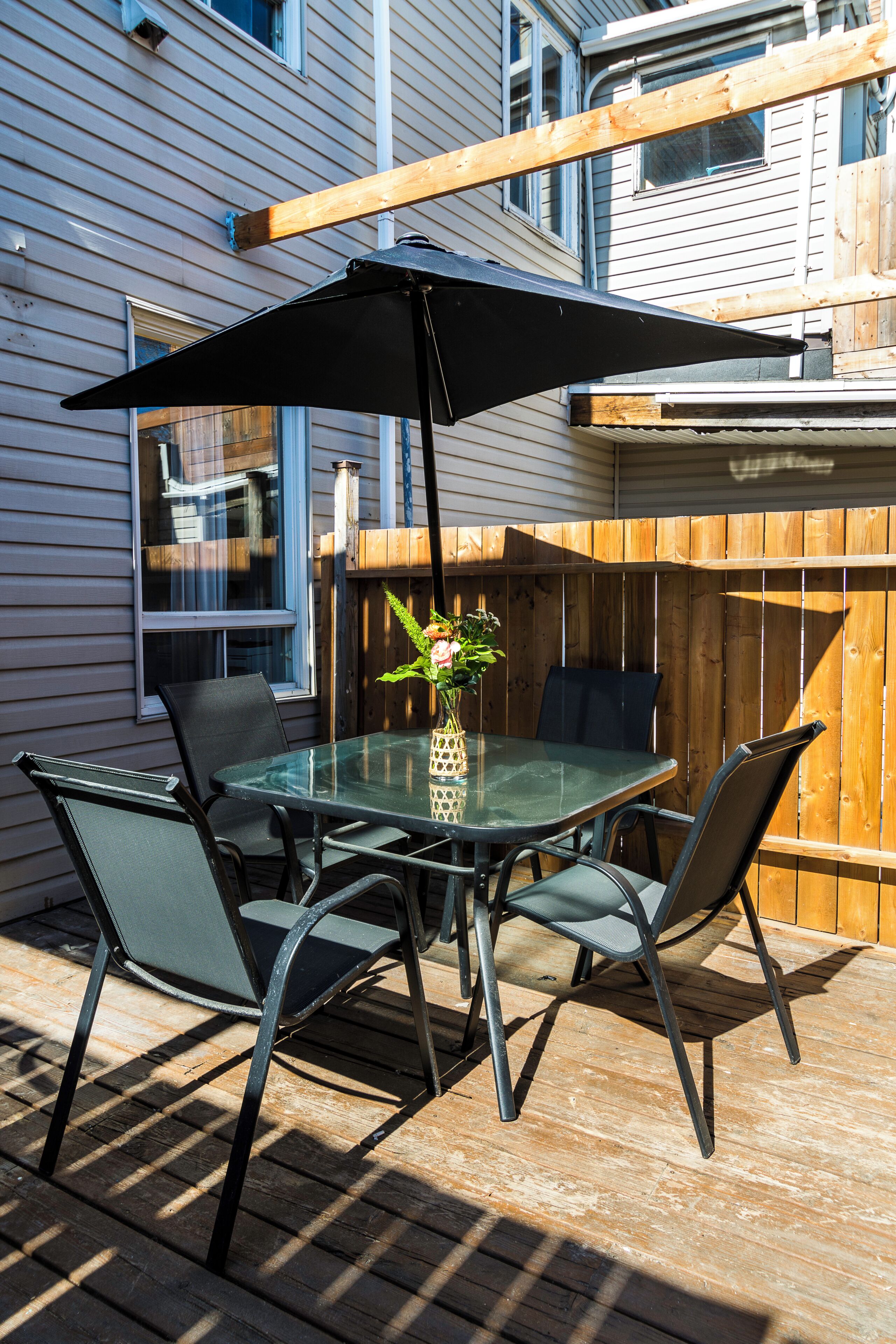 An outdoor patio with a glass table, four chairs, a black umbrella, and a vase with flowers.