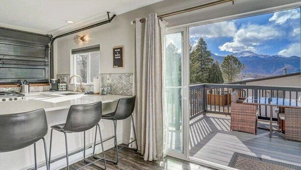 A modern kitchen with a bar area, a dining space with chairs, and a balcony with a view of mountains. - PeakView Lil Lux! Hot Tub & Mountain Views (Colorado Springs)